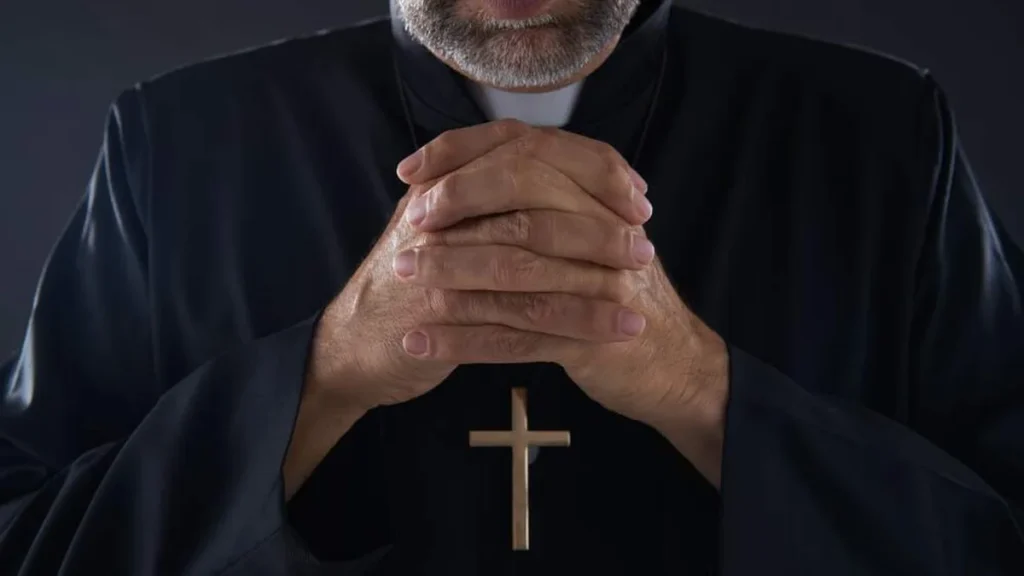 Imagem padre vestindo tunica preta e crucifixo de madeira rezando de maos postas do artigo Por que os padres modernos usam cada vez menos as vestes clericais?