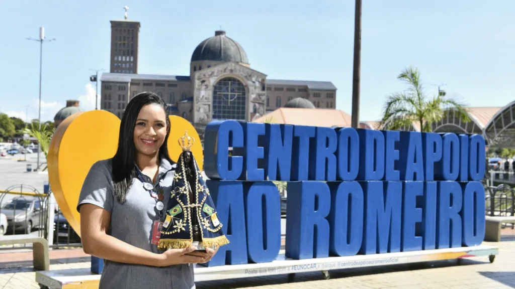 Imagem mulher a frente do letreiro do artigo Centro de Apoio ao Romeiro: comprometido com o devoto da Mãe Aparecida