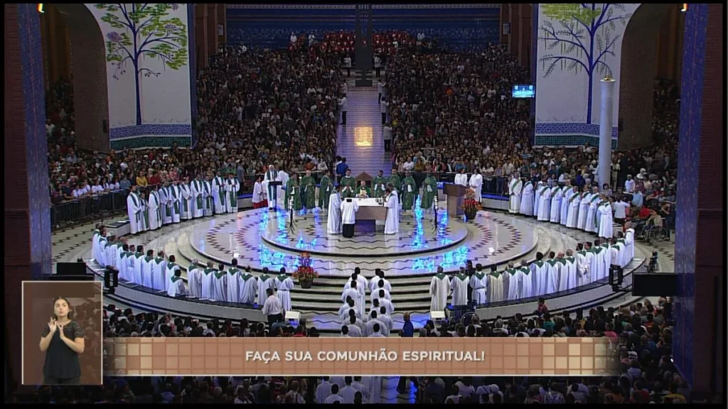 Foto panorâmica do altar central dentro do Santuário Nacional de Aparecida. Dezenas de bispos e padres estão no presbitério. Centenas de devotos estão em pé ao redor do altar. Na tela, a janela da intérprete de Libras e a tarja "Faça sua comunhão esp