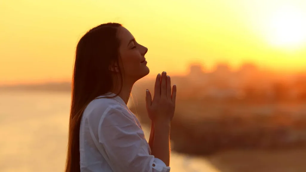 Imagem mulher rezando com o por do sol do artigo Preciso rezar em voz alta para que Deus me escute?