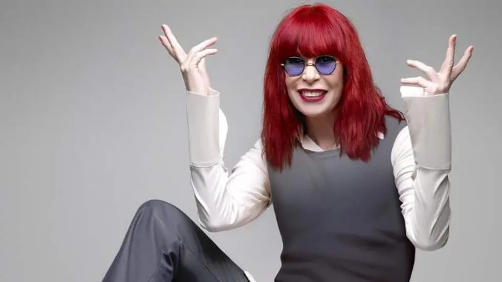 Rita Lee com cabelo vermelho e óculos, roupa cinza e branca fazendo pose com mãos pra cima