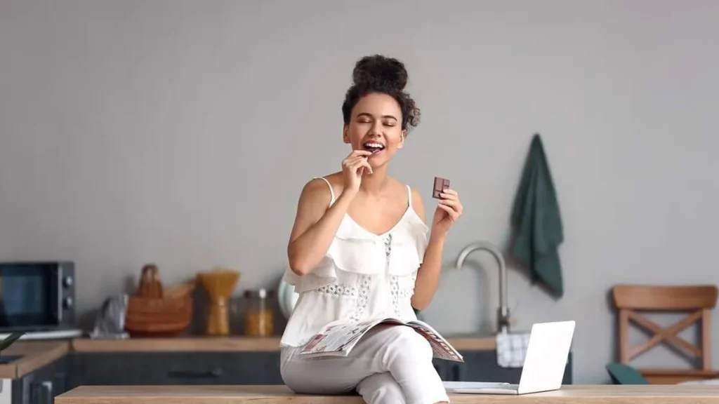 Moça sentada na bancada da cozinha comendo chocolate