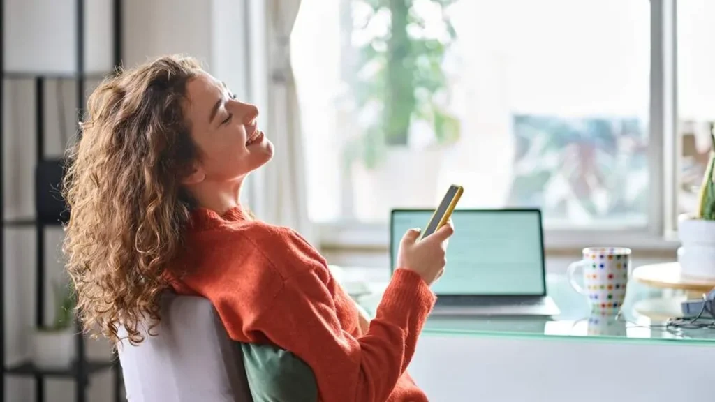 Imagem mulher sorrindo tranquila e sentada com o celular na mao do artigo Como a "Santa Indiferença" fortalece a nossa fé?