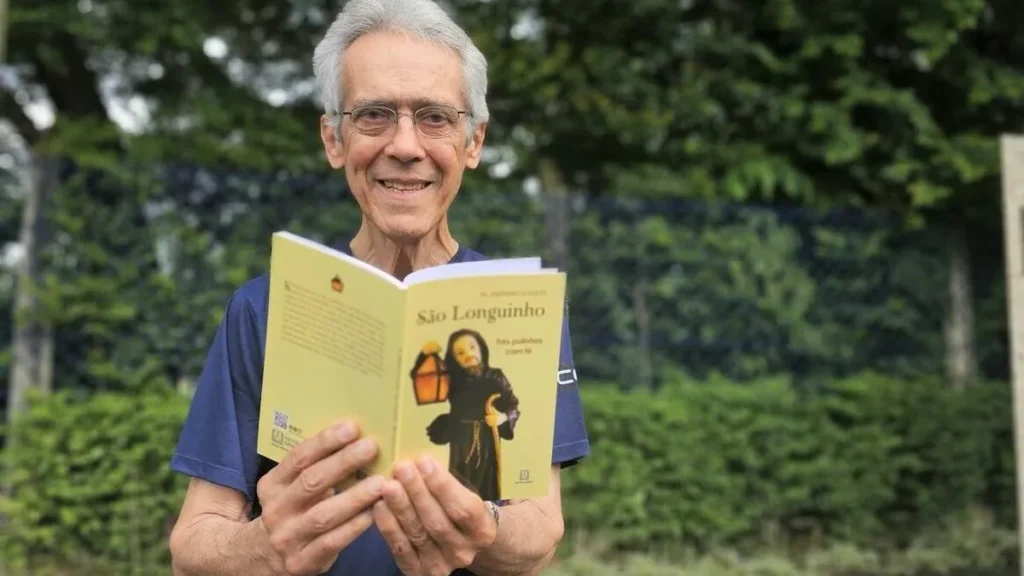 Imagem Pe. Domingos Savio com Livro de Sao Longuinho do artigo "Três pulinhos com fé" retrata a missão de São Longuinho