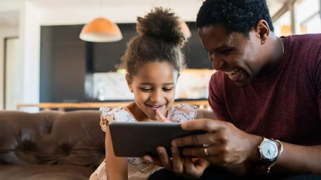 Imagem retrato de uma filha e um pai se divertindo juntos e brincando com um tablet digital em casa do artigo Adultização e a necessária atenção da família