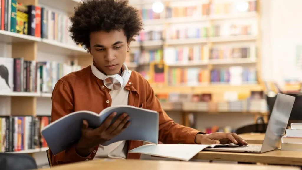 Imagem garoto negro estudando em uma biblioteca com notebook e livros sobre a mesa do artigo 5 livros essenciais para universitários lerem durante o curso