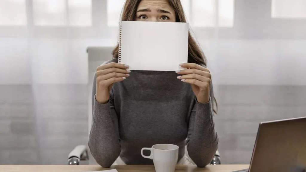 Imagem empresaria cobrindo o rosto com um bloco de notas do artigo Síndrome do impostor impacta carreira de muitos profissionais