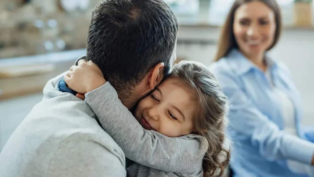 Imagem menina carinhosa com os olhos fechados abracando o pai em casa mae esta ao fundo Easy Resize.com do artigo Um convite da vida: redescobrir a essência na vida cotidiana