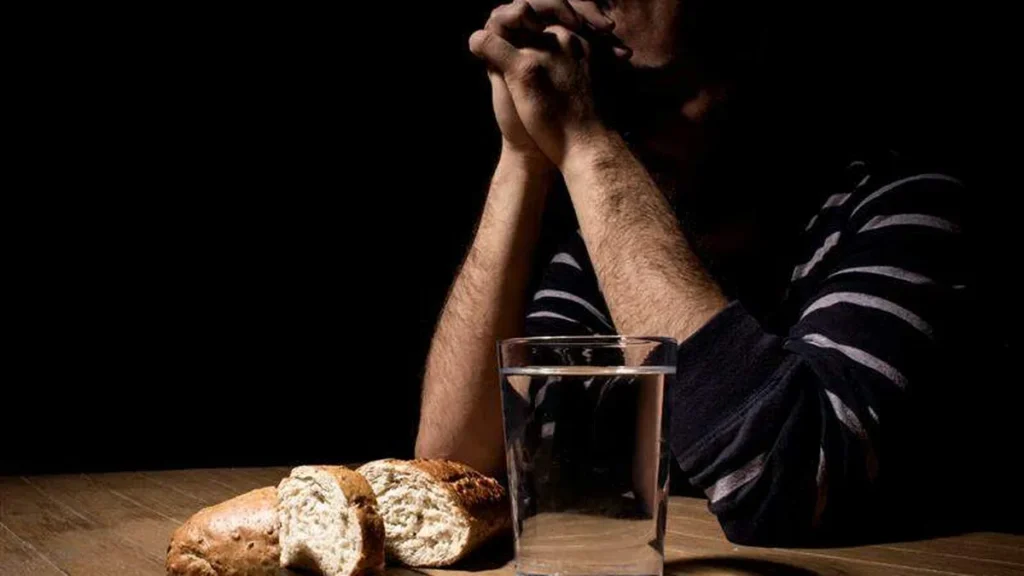 Imagem Homem com as maos unidas em oracao sob a mesa com um copo de agua e um pao do artigo Mistérios da Fé: entenda o jejum e penitência na modernidade